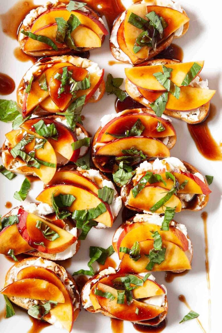 Sliced baguette topped with goat cheese, peach slices, chopped basil, and a drizzle of balsamic glaze arranged on a white platter to make peach crostini at home.