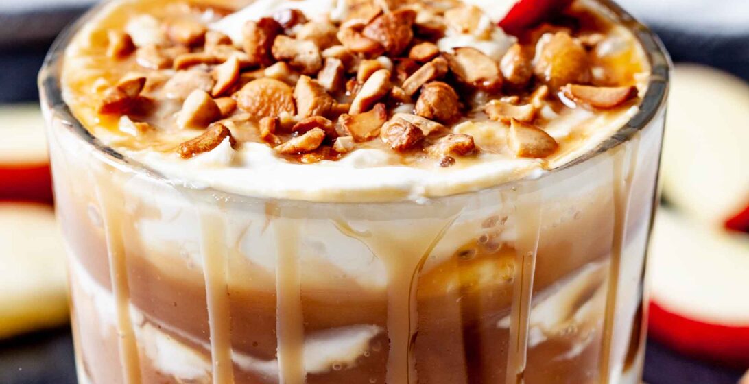 A glass filled with creamy caramel apple cheesecake dip is layered with cream, topped with chopped nuts, a caramel drizzle, and a slice of red apple.