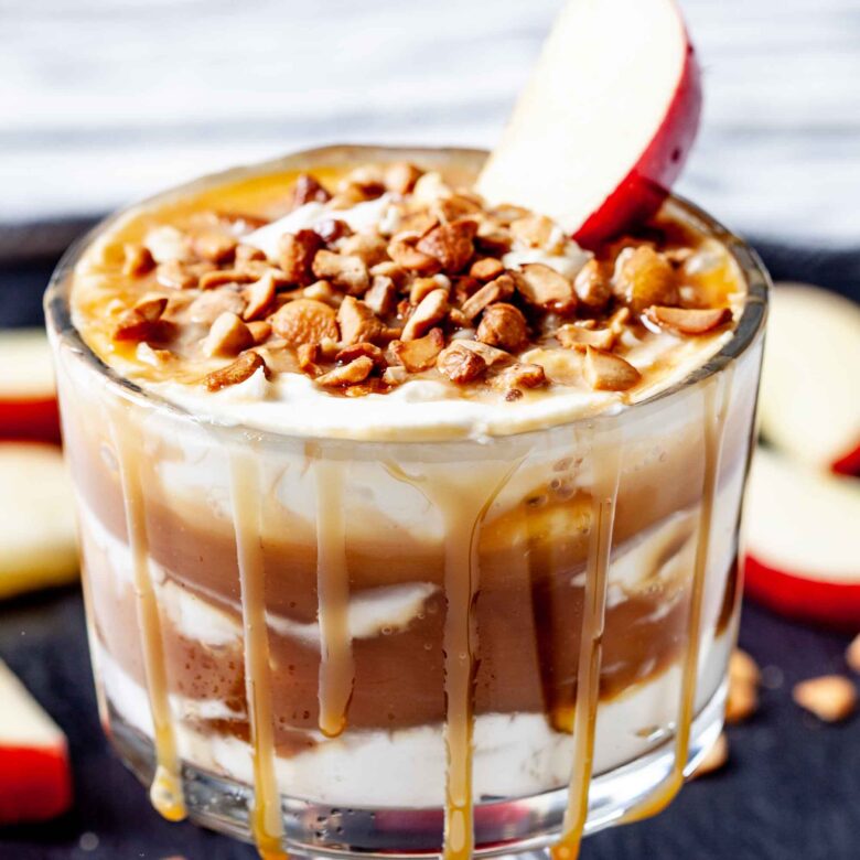 A glass filled with creamy caramel apple cheesecake dip is layered with cream, topped with chopped nuts, a caramel drizzle, and a slice of red apple.