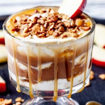 A glass filled with creamy caramel apple cheesecake dip is layered with cream, topped with chopped nuts, a caramel drizzle, and a slice of red apple.