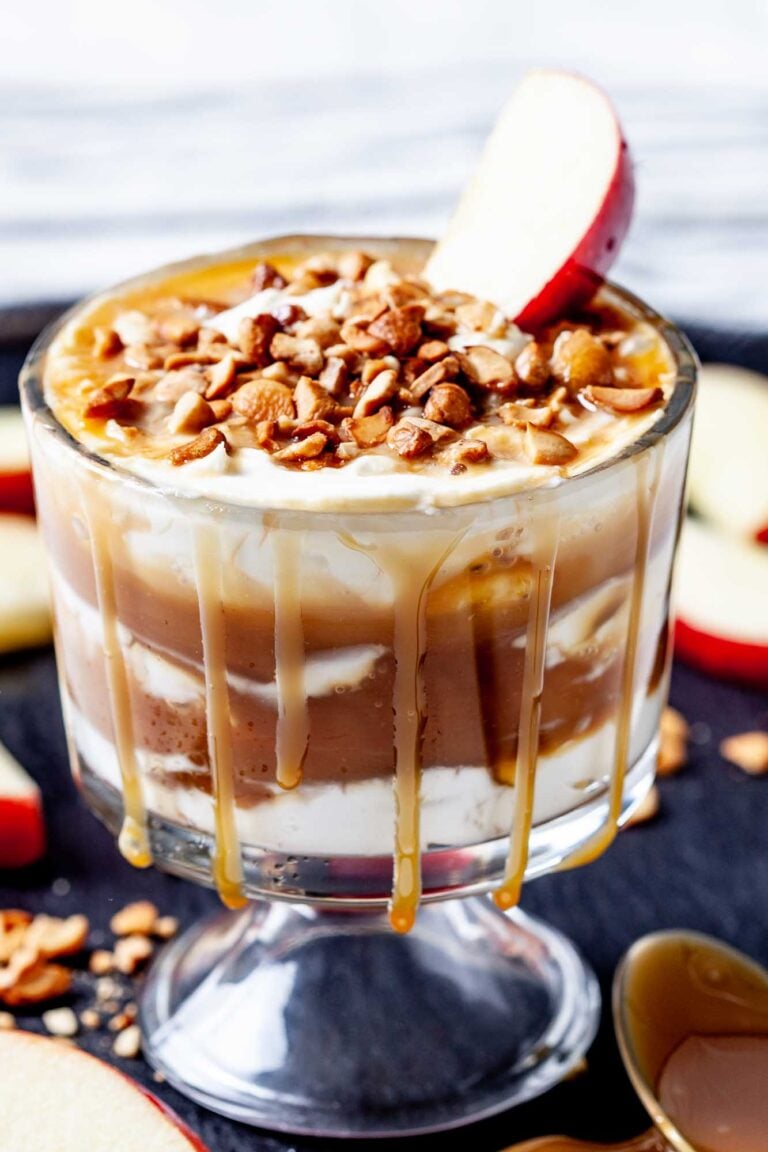 A glass filled with creamy caramel apple cheesecake dip is layered with cream, topped with chopped nuts, a caramel drizzle, and a slice of red apple.
