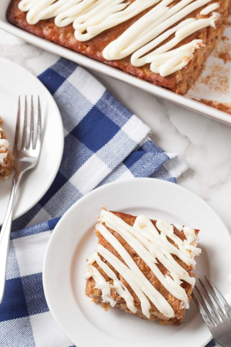 A square piece of easy apple cake with white icing is served on a plate next to a fork, with a baking pan of more cake and a blue checkered napkin nearby.