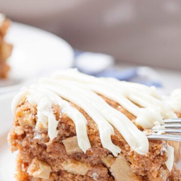 A close-up of a slice of easy apple cake with cream cheese icing drizzled on top, served on a white plate with a fork.
