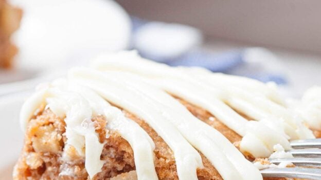 A close-up of a slice of easy apple cake with cream cheese icing drizzled on top, served on a white plate with a fork.