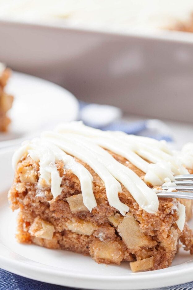 A close-up of a slice of easy apple cake with cream cheese icing drizzled on top, served on a white plate with a fork.