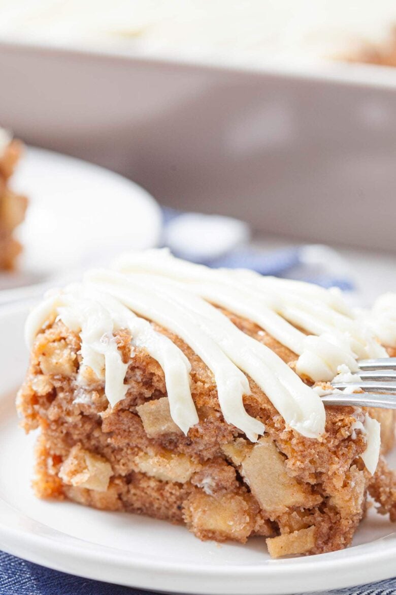 A close-up of a slice of easy apple cake with cream cheese icing drizzled on top, served on a white plate with a fork.