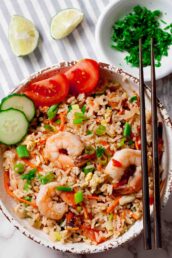 A bowl of Thai fried rice with garlic shrimp, sliced cucumber, cherry tomatoes, green onions, and chopsticks; lime wedges and chopped herbs on the side.