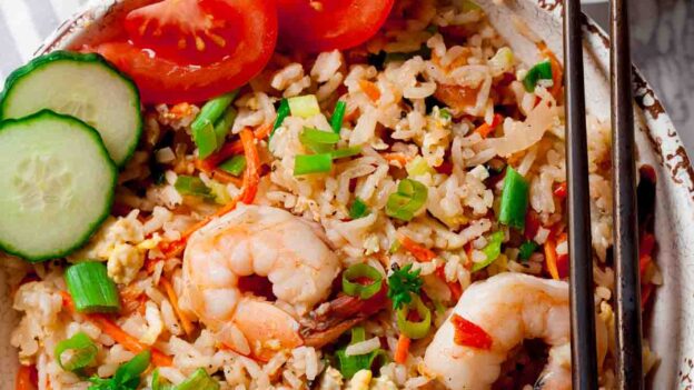 A bowl of Thai fried rice with garlic shrimp, sliced cucumber, cherry tomatoes, green onions, and chopsticks; lime wedges and chopped herbs on the side.