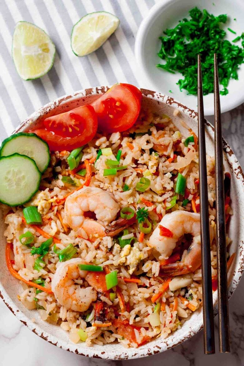 A bowl of Thai fried rice with garlic shrimp, sliced cucumber, cherry tomatoes, green onions, and chopsticks; lime wedges and chopped herbs on the side.