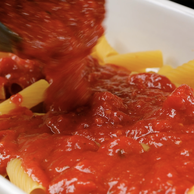 Marinara being poured over uncooked rigatoni in a white baking dish.