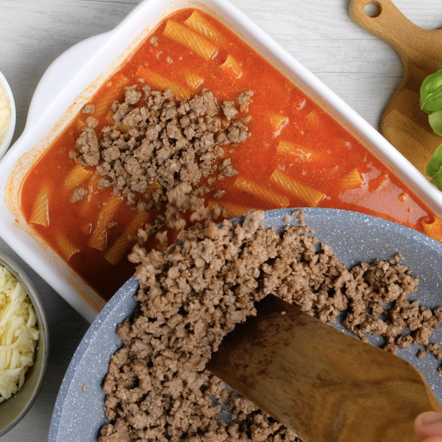 Cooked sausage being layered on top of baked rigatoni mixture in a white casserole.