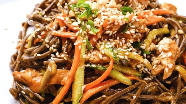 Peanut soba noodles with chicken, served with lime wedges on the side and a sesame seeds on top.