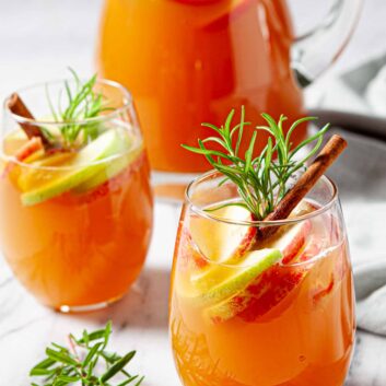 Two glasses of orange-colored Apple Cider Sangria with apple and lime slices, cinnamon sticks, and rosemary sprigs.
