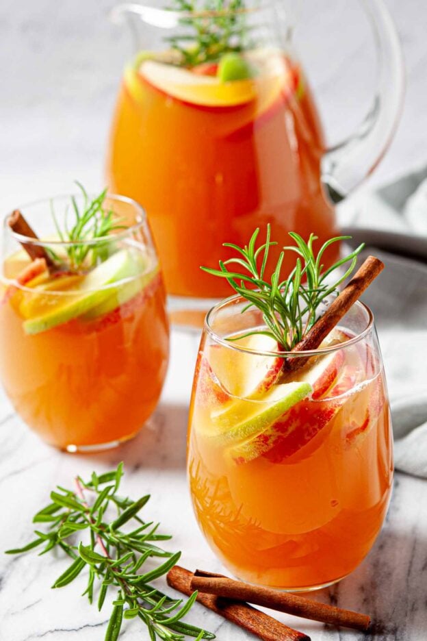 Two glasses of orange-colored Apple Cider Sangria with apple and lime slices, cinnamon sticks, and rosemary sprigs.