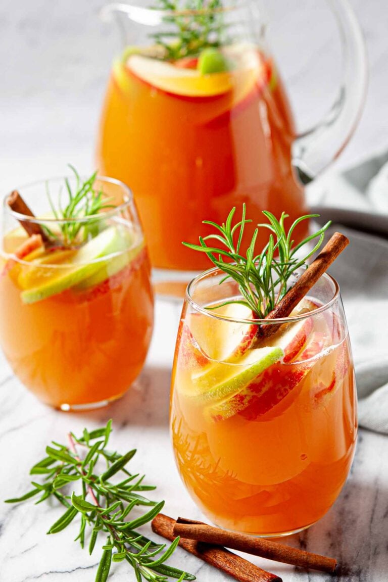 Two glasses of orange-colored Apple Cider Sangria with apple and lime slices, cinnamon sticks, and rosemary sprigs.
