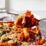 A close-up of baked rigatoni with savory sausage, marinara, and melted cheese being served from a casserole dish with a spoon.
