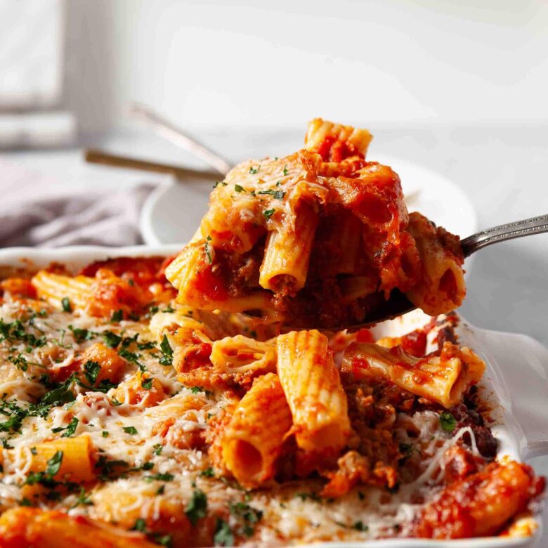 A close-up of baked rigatoni with savory sausage, marinara, and melted cheese being served from a casserole dish with a spoon.