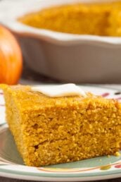 A slice of pumpkin cornbread topped with a pat of butter sits on a plate, with a baking dish and a pumpkin in the background.