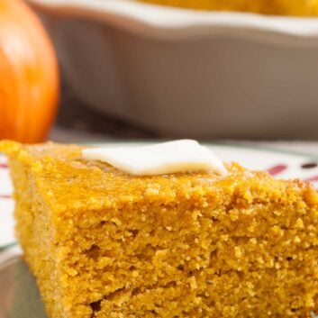 A slice of pumpkin cornbread topped with a pat of butter sits on a plate, with a baking dish and a pumpkin in the background.