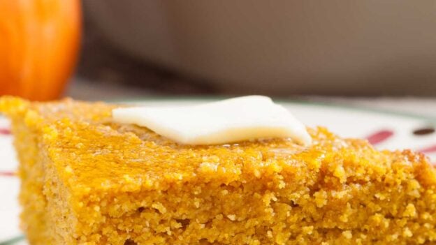 A slice of pumpkin cornbread topped with a pat of butter sits on a plate, with a baking dish and a pumpkin in the background.