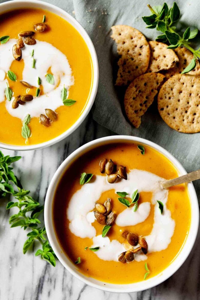 Overhead shot of two bowls with curry butternut squash soup, with coconut milk on top, and garnished with fresh herbs and roasted pepitas.