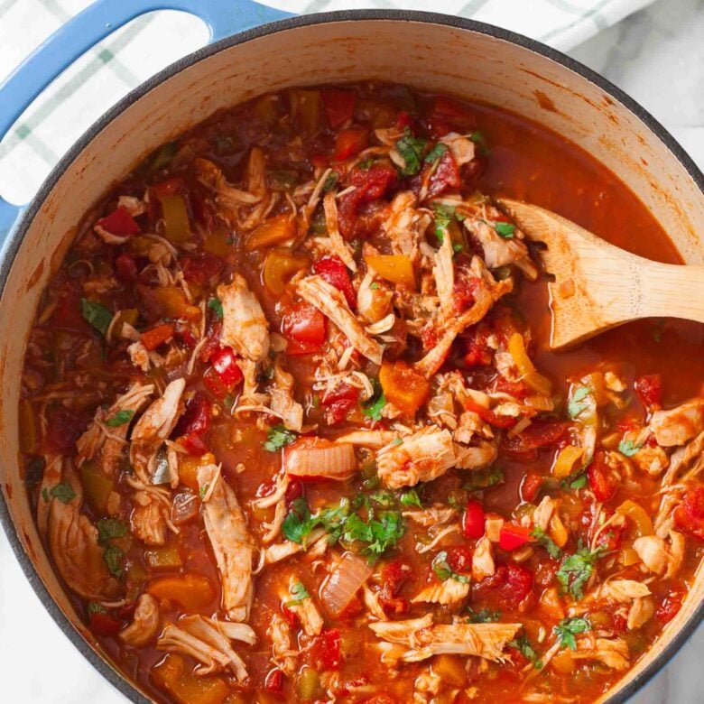 A pot of healthy Chicken Chili with tomatoes, onions, peppers, and herbs, being stirred with a wooden spoon.