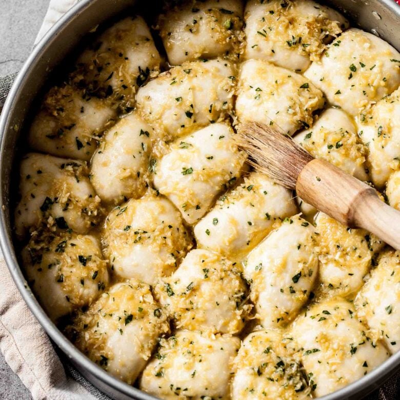 A pastry brush brushing garlic parmesan butter mixture of dough rolls in a pan.