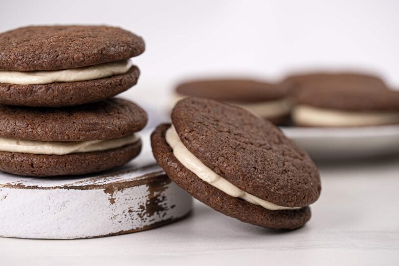 Peanut butter fudge cookies are stacked and arranged on a wooden surface, with more sandwich cookies on a plate in the background.
