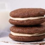 Two chocolate sandwich cookies with creamy filling are stacked on a white surface, with a blurred glass of milk in the background.
