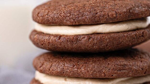 Two chocolate sandwich cookies with creamy filling are stacked on a white surface, with a blurred glass of milk in the background.