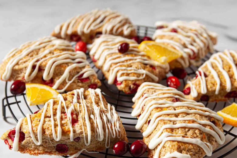 Cranberry orange scones drizzled with melted chocolate on a cooling rack with fresh cranberries and orange slices on the side.