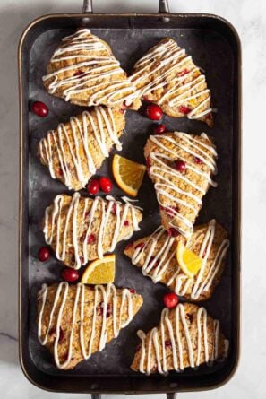 A baking tray with eight Cranberry Orange Scones drizzled with white icing, garnished with fresh cranberries and citrusy orange slices on parchment paper.