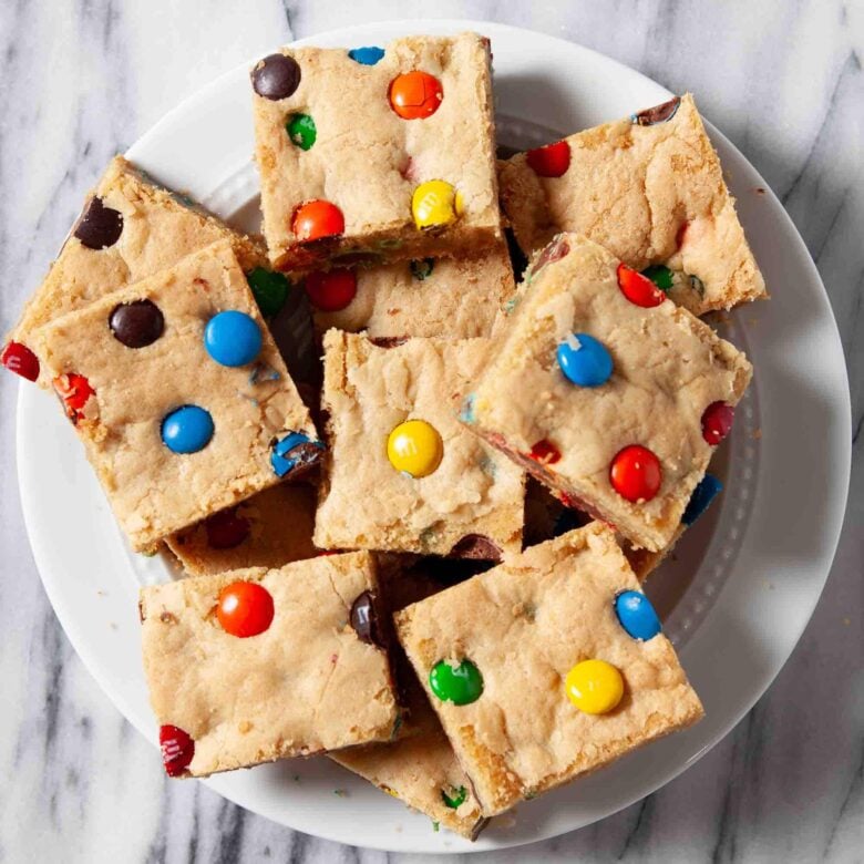 A white plate holding several chewy M&M cookie bars on a marble surface.