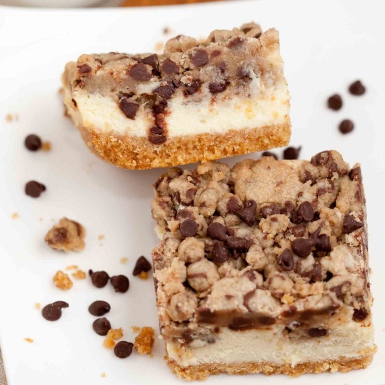 Two Chocolate Chip Cheesecake Bars with a creamy filling and chocolate chip cookie crumble topping sit on a white plate, surrounded by scattered chocolate chips and crumbs.