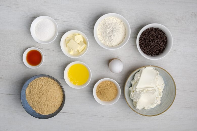 Ingredients to make chocolate chip cheesecake bars on a light surface.