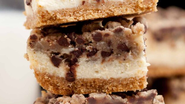 Three stacked Chocolate Chip Cheesecake Bars with a graham crust, creamy filling, and cookie topping are photographed close-up.