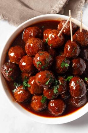 A white bowl filled with grape jelly meatballs in a dark glaze, garnished with chopped herbs, with two toothpicks inserted.