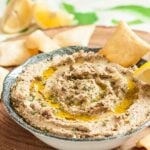 A bowl of lentil hummus drizzled with olive oil, garnished with herbs, and served with pita chips and lemon wedges on a wooden surface.