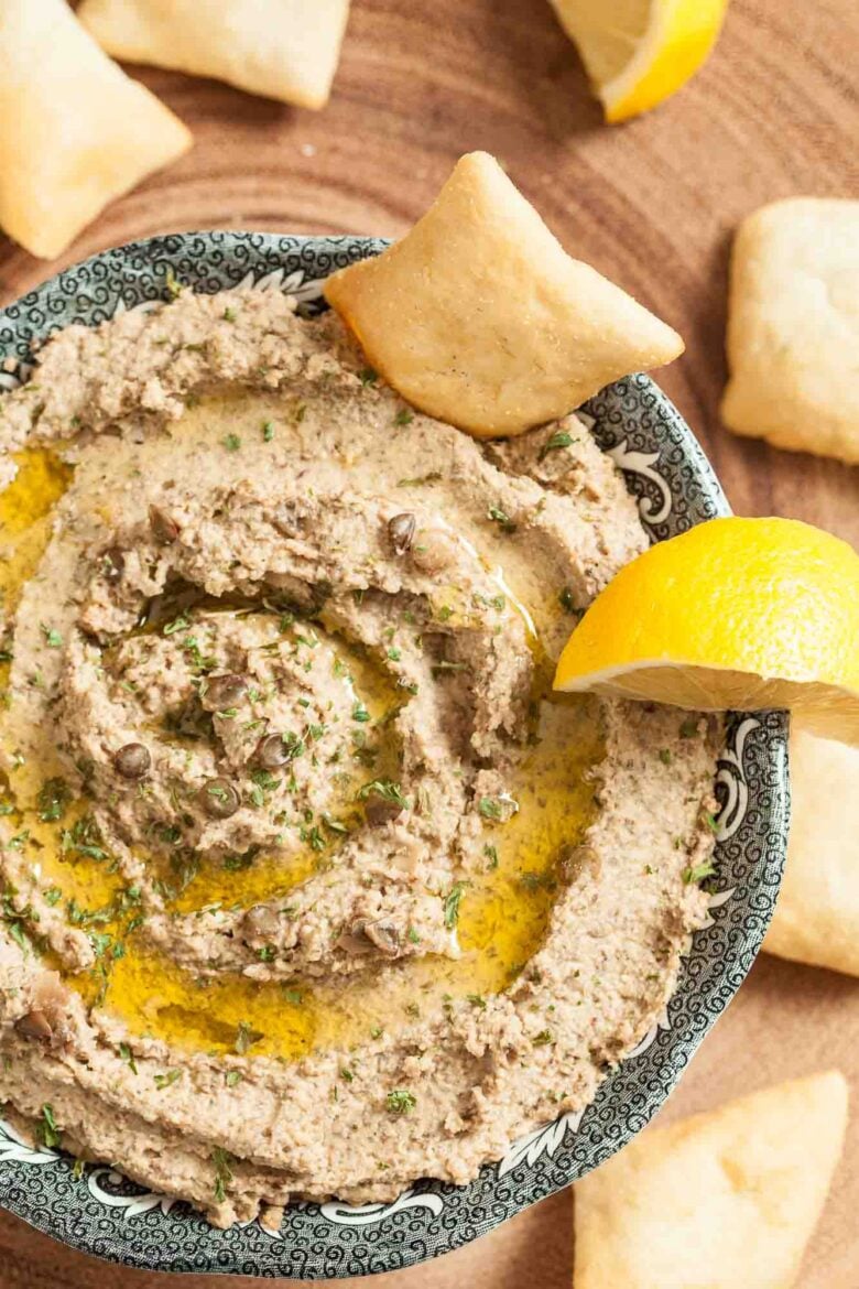 A bowl of lentil hummus garnished with herbs, capers, and olive oil, served with pita chips and a lemon wedge on the side.