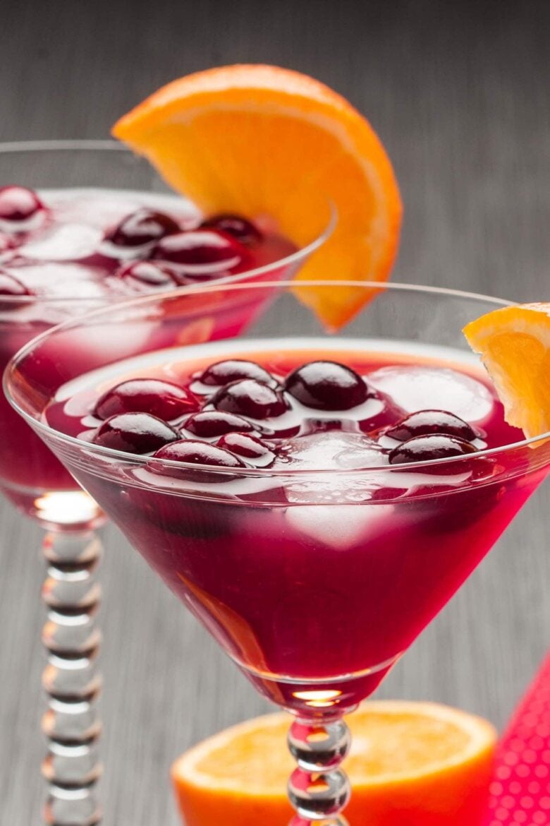 Two martini glasses filled with a vibrant purple Madras cocktail, ice, and berries, each garnished with an orange slice.