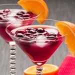 Two martini glasses filled with a vibrant purple Madras cocktail, ice, and berries, each garnished with an orange slice.
