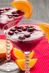 Two martini glasses filled with a vibrant purple Madras cocktail, ice, and berries, each garnished with an orange slice.
