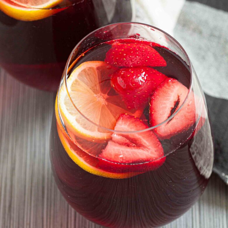 A glass of pomegranate punch, features sliced strawberries and lemon floating on top and is set on a striped surface.