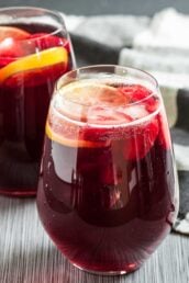 A glass of pomegranate punch with ice cubes and citrus slices, made with simple ingredients, sits on a striped surface, with another glass in the background.