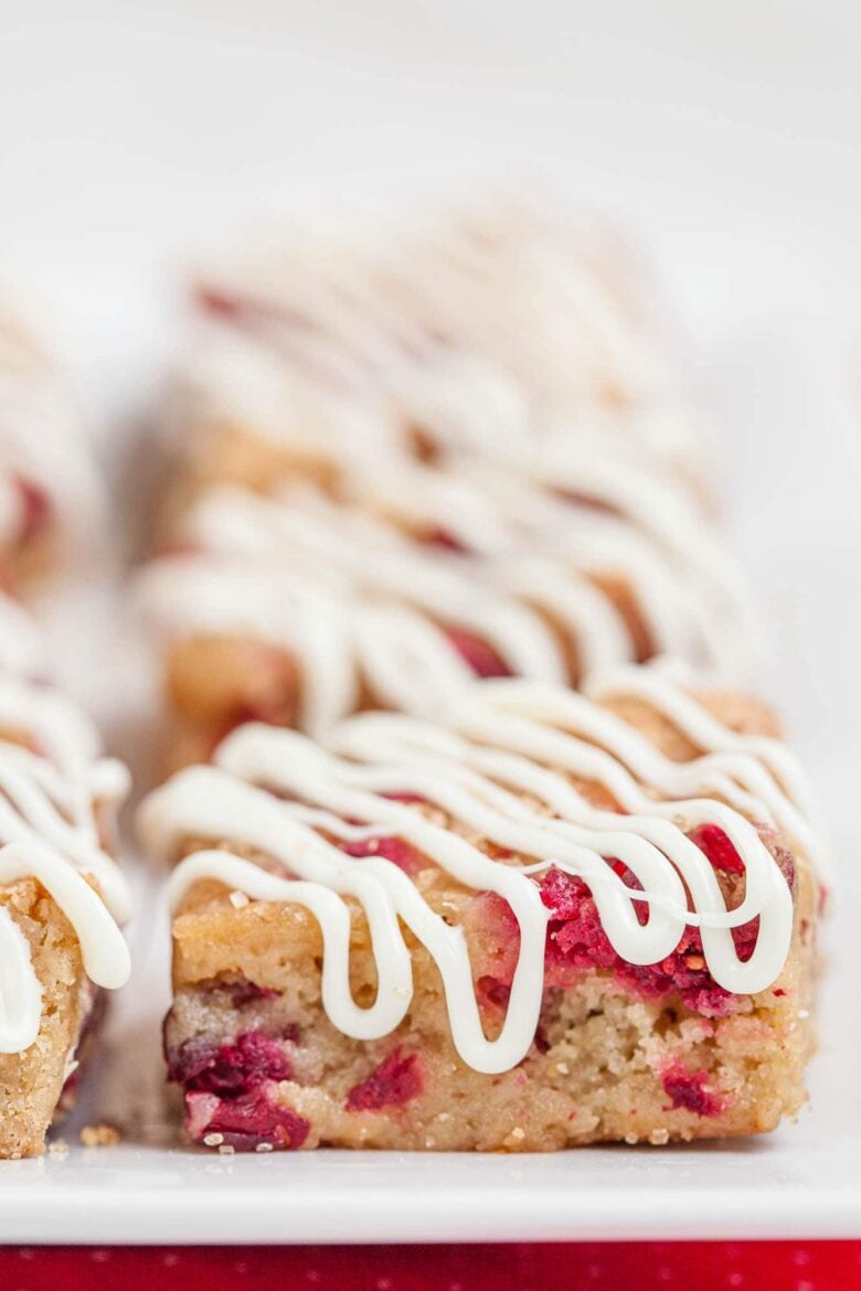 Close-up of Chewy White Chocolate Cranberry Bars with nuts and fresh cranberries, topped with a drizzle of white icing, arranged in rows on a white surface.