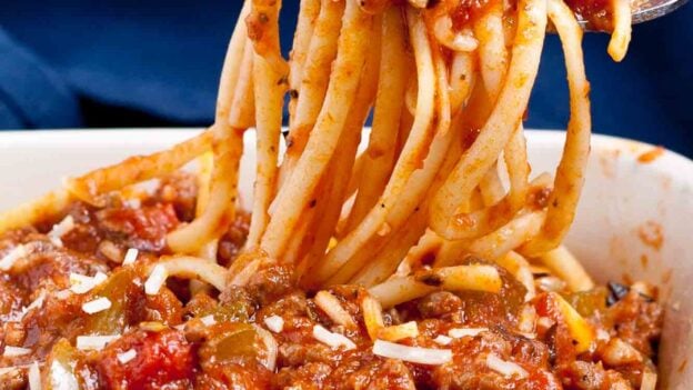 A fork lifts a serving of Italian sausage pasta, topped with thick meat and tomato sauce, from a red dish.
