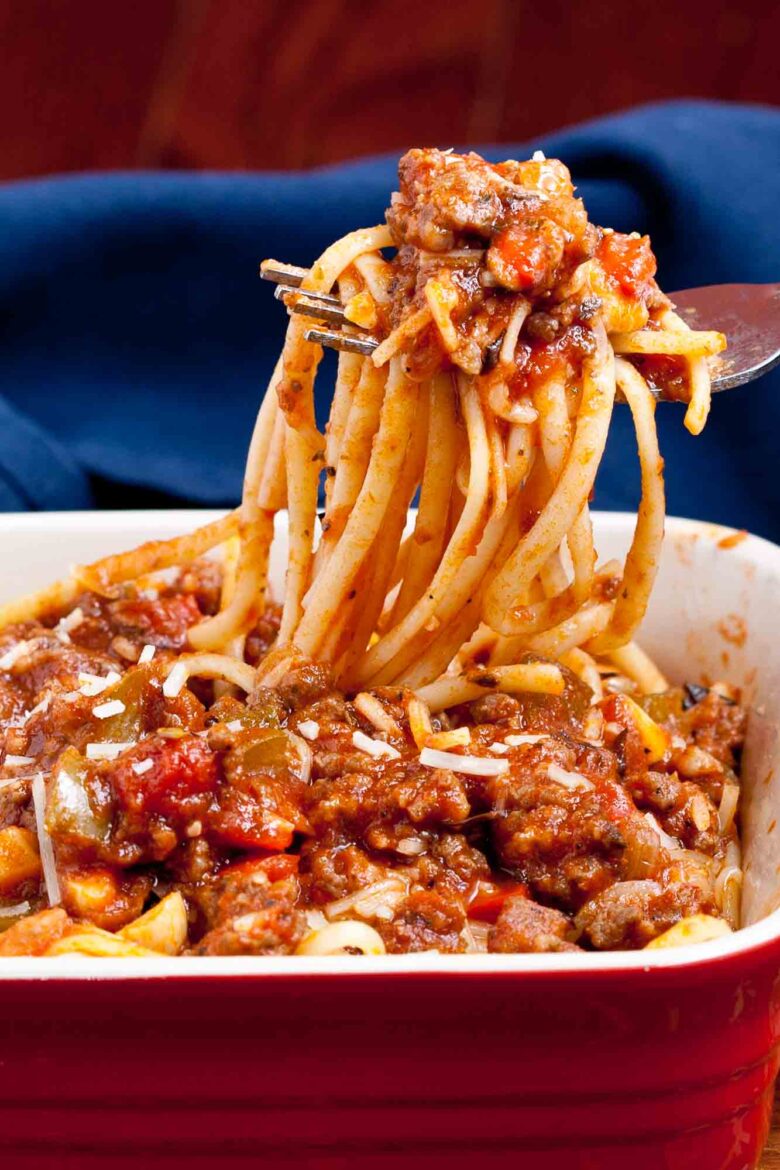 A fork lifts a serving of Italian sausage pasta, topped with thick meat and tomato sauce, from a red dish.