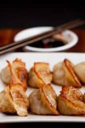 Closeup shot of potsticker on a white plate with dipping sauce and chopsticks in the background.