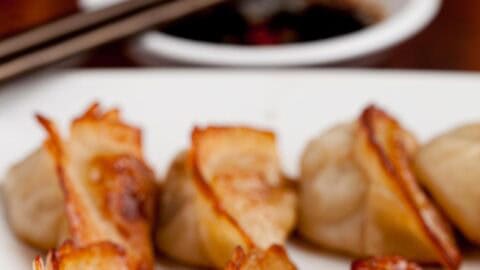 Closeup shot of potsticker on a white plate with dipping sauce and chopsticks in the background.