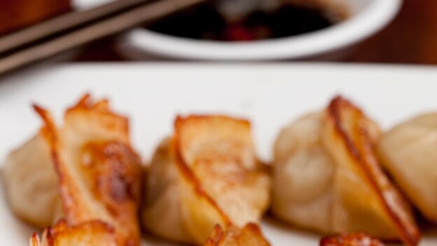 Closeup shot of potsticker on a white plate with dipping sauce and chopsticks in the background.